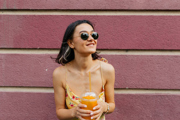 ピンクの壁にオレンジのカクテルでポーズを取った夏のドレスを身に着けている短い髪型の魅力的な笑顔の女性。高品質の写真 - 写真・画像