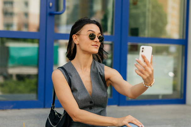 Κομψή μοντέρνα γυναίκα με κοντά μαλλιά φορώντας γυαλιά ηλίου κάνει selfie, ενώ το περπάτημα στο δρόμο. Υψηλής ποιότητας φωτογραφία - Φωτογραφία, εικόνα