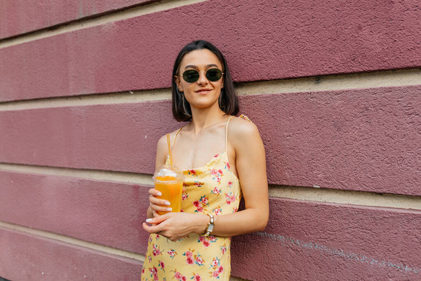 Suloinen kaunis moderni tyttö kesällä kirkas mekko tilalla smoothie poseeraa vaaleanpunainen seinä ja nauttia viikonloppuna. Laadukas kuva - Valokuva, kuva