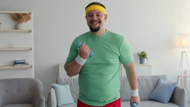 Happy overweight man enjoying fitness activity at home, exercising with dumbbells and smiling to camera - Záběry, video