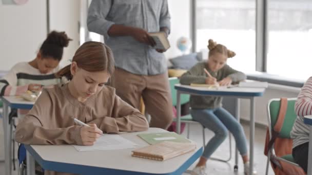 Mittlere Aufnahme eines smarten 12-jährigen kaukasischen Schulmädchens, das am Schreibtisch im Klassenzimmer sitzt und Test als männlicher afroamerikanischer Lehrer macht, der ihr bei harten Übungen hilft - Filmmaterial, Video