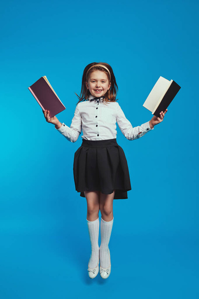 Schoolgirl holds two notebooks while jumping high over blue background - Foto, Bild
