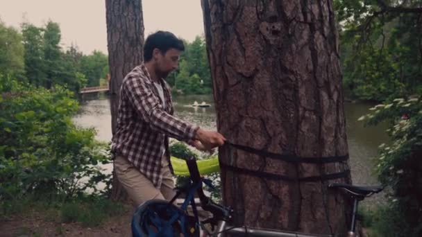 Männliche Touristen kamen mit dem Fahrrad auf dem Campingplatz in der Nähe des Sees an. Radler stellen Hängematte im Wald am Fluss auf. Reisende binden Hängematte im Park an Teich. Guy befestigt Hängemattengurt an Baum - Filmmaterial, Video