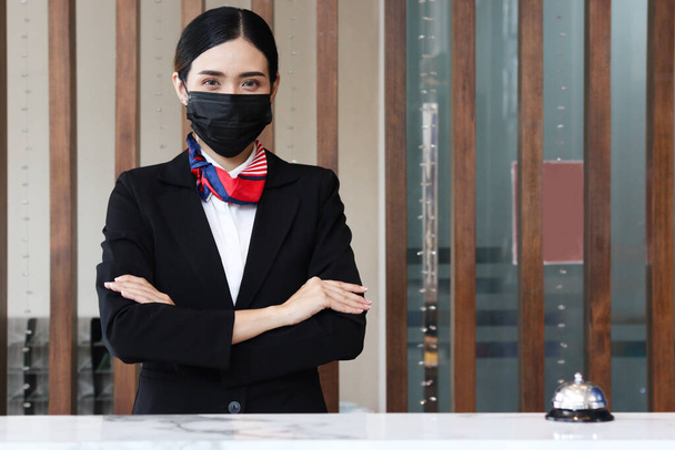 Portrait of young beautiful Asian female receptionistin suit wearing face mask to prevent corona virus infection, confidential standing with arms crossed at hotel reception counter desk, check in hotel service on vacation concept.   - Φωτογραφία, εικόνα