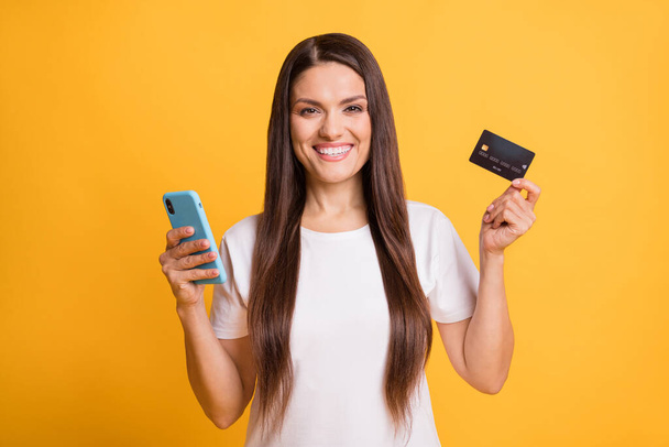 Photo portrait of happy woman smiling using mobile phone keeping plastic bank card isolated on vibrant yellow color background - Zdjęcie, obraz