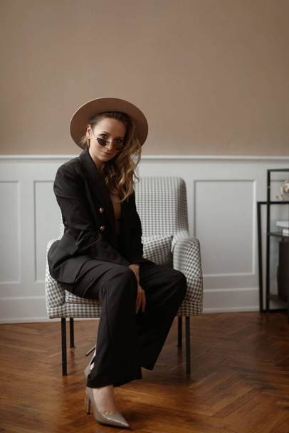 Young model woman with gentle makeup wearing trendy hat and modish sunglasses sitting in a vintage armchair at the classic interior - Fotoğraf, Görsel