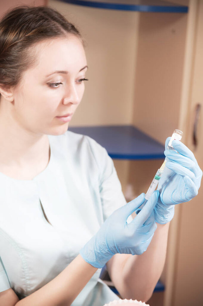A young doctor Caucasian woman in medical clothes and rubber gloves without a protective mask is drawing a vaccine or medicine from an ampoule into a syringe. Failure to comply with medical safety - Foto, afbeelding