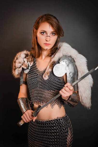 Medieval woman warrior in chain mail armor and polar fox fur on her shoulders standing with a sword in hand on a dark background. Fantasy barbarian, viking. - Foto, afbeelding