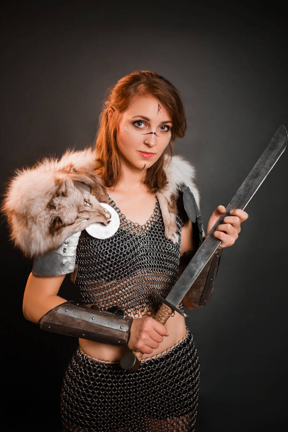 Medieval woman warrior in chain mail armor and polar fox fur on her shoulders standing with a sword in hand on a dark background. Fantasy barbarian, viking. - Fotografie, Obrázek