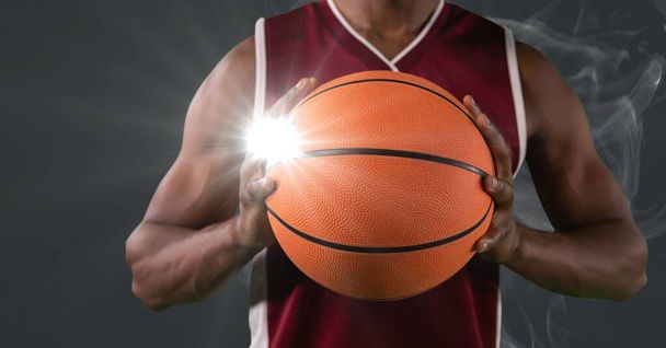 Mid section of male basketball player holding ball against smoke and light spot on grey background. sports tournament and competition concept - Foto, Imagem