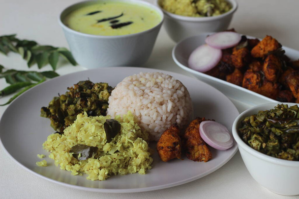 Ei-kasvisruoat valmistetaan Kerala tyyliin. Tarjoilu sisältää keitettyä punaista riisiä, sekoita paistettua sipulia raastetun kookospähkinän kanssa, sekoita paistettua okraa, karkaistua kirnupiimää, poppadumia ja ilmaa paistettua basakalaa.. - Valokuva, kuva