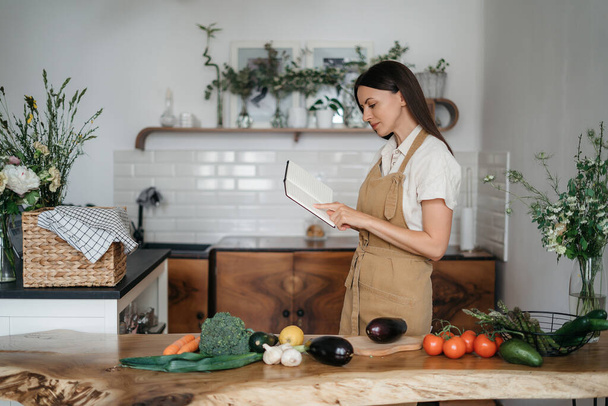 beautiful young brunette woman prepares a healthy meal from fresh organic vegetables in the kitchen according to a recipe from a notebook - Foto, imagen
