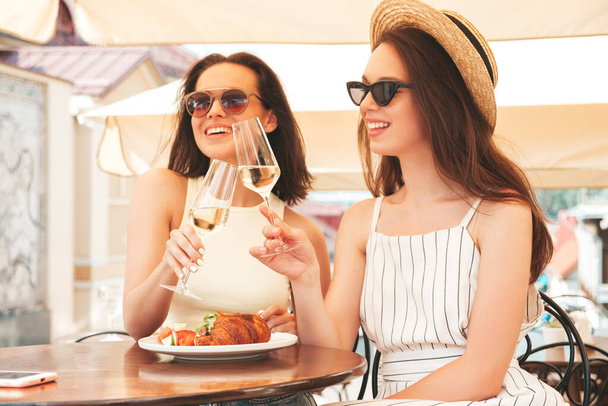 Two young beautiful smiling hipster female in trendy summer clothes.Carefree women posing at veranda cafe in the street.Positive models drinking white vine.Eating - Foto, Imagen