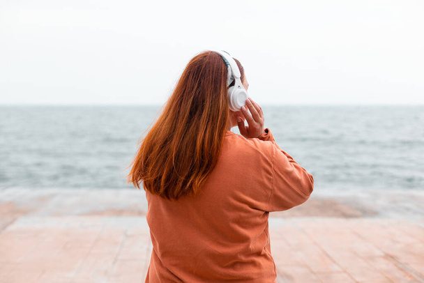 задний вид портрета молодой девушки медитирует с беспроводными наушниками на открытом воздухе на пляже с видом на море - Фото, изображение