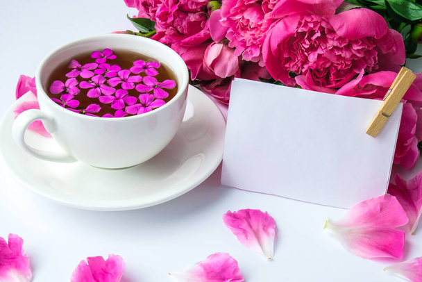 Kreatives Layout mit rosa Pfingstrosen-Blüten und einer Tasse Tee auf hellem Tisch. Papiernotiz. Kopierraum. Frühling Valentinstag, Frau, Mutter, Urlaub am 8. März, Romantikfrühstück. Stilvolle Bloggerin. Grußkarte vorhanden. Einladung. Blog-Eintrag - Foto, Bild