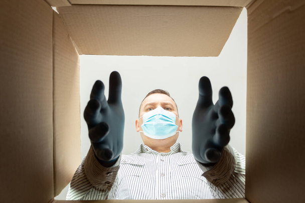 Egy férfi orvosi maszkban és kesztyűben kivesz valamit a dobozból. Lényegében. Vegyél ki valamit egy kartondobozból. - Fotó, kép