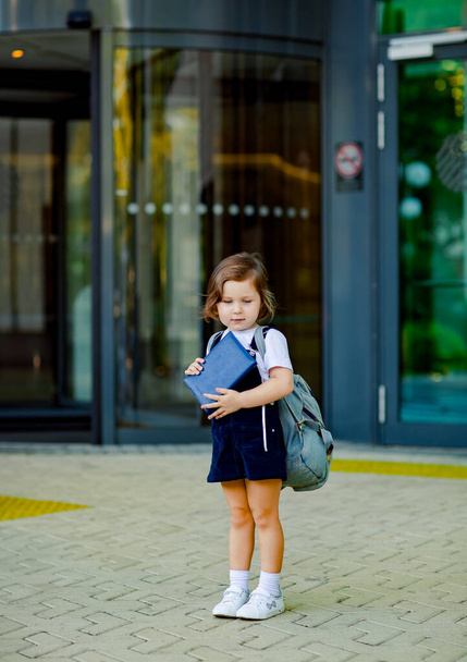 a beautiful Caucasian girl, a schoolgirl, is standing near the school, with a backpack and holding a diary in her hands - Photo, image