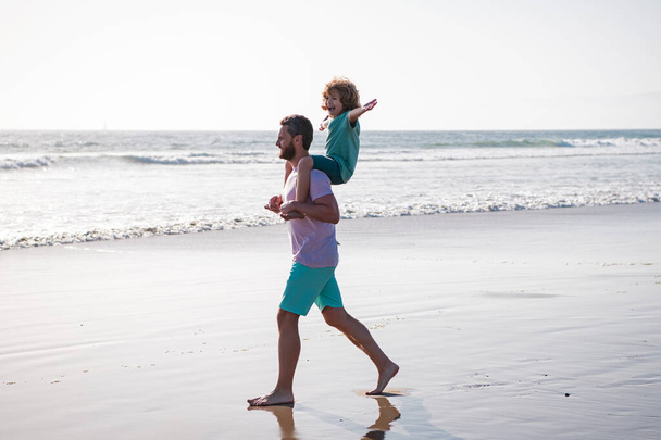 Father and son walking on sea. Handsome man father carrying young boy son. Happy dad holding child. Daddy with kid boy on a summer day. Concept of friendly family and of summer vacation. - Foto, afbeelding