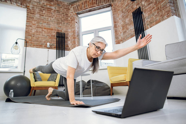 Retired woman using laptop and doing yoga at home - Fotoğraf, Görsel
