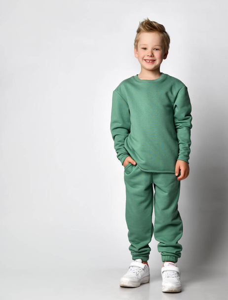 aranyos aktív kisfiú divatos sport zöld ruhák álló kéz a zsebben mutatja érzelmek a boldogság és a kényelem. - Fotó, kép