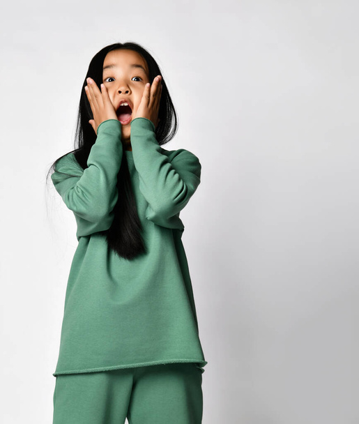 Shocked surprised asian preteen girl in green clothing touching cheeks - Foto, imagen
