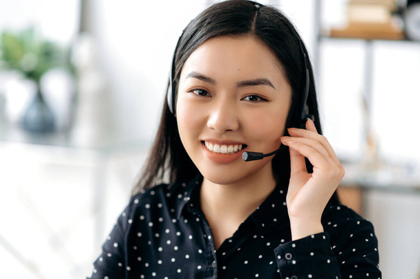 Közeli kép egy kellemes, magabiztos, hosszú hajú, fiatal ázsiai nő headset, szabadúszó, call center munkavállaló vagy tanácsadó, visel fekete elegáns ing, néz közvetlenül a kamera, mosolyog barátságos - Fotó, kép
