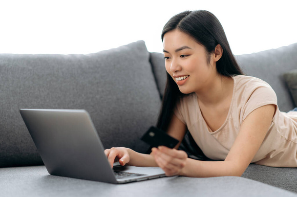 Online shopping. Positive beautiful young asian brunette woman lying on a couch, uses laptop and credit card to pay for online purchases and delivery, enters data, looks at the screen, smiles - Photo, Image