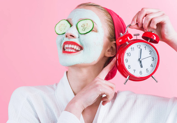 Smiling Woman with cosmetic facial mask and alarm clock in hand. Cosmetics procedure. Beauty spa and cosmetology. - Fotó, kép