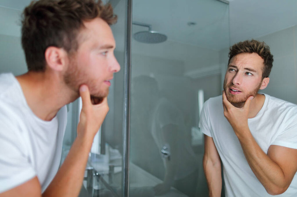 Man face care looking in mirror touching his beard or skin health. Male beauty morning skincare routine in home bathroom. After shave men lifestyle concept - Fotoğraf, Görsel