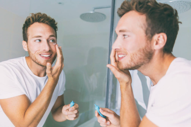 Man putting skincare facial treatment cream on face. Anti-aging skin care product. Male beauty morning routine at home lifestyle. Guy looking in bathroom mirror applying moisturizer under eyes - Fotoğraf, Görsel