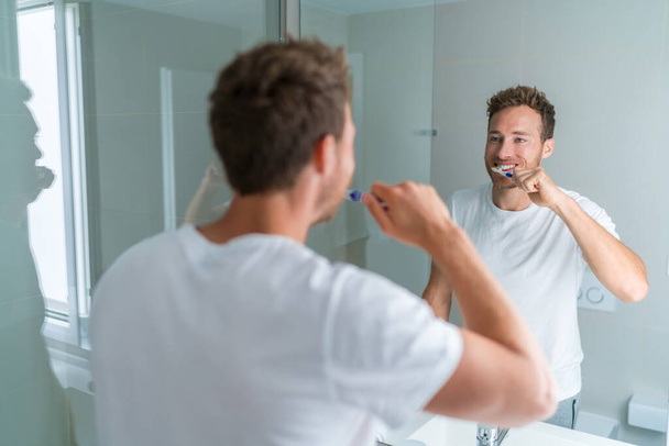 Man getting ready in the morning doing hygiene routine brushing his teeth looking in mirror of home bathroom using toothbrush in for clean dental oral care - Foto, Imagem