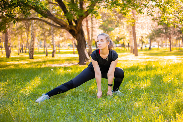 A young, slender woman in a sports uniform does a side lunge exercise to train the muscles of the legs. Fitness classes in nature.  The concept of independent sport training in park. - Photo, Image