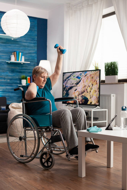 Mujer mayor no válida en silla de ruedas viendo gimnasio ejercicio corporal en la tableta en la sala de estar - Foto, Imagen