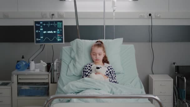 Πορτρέτο νοσηλευόμενου ασθενή με άρρωστο παιδί που κρατά αρκουδάκι που αναπαύεται στο κρεβάτι κατά τη διάρκεια ιατρικής γνωμάτευσης - Πλάνα, βίντεο