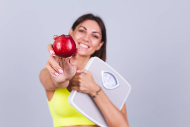 Femme sportive se tient sur fond gris, satisfait des résultats de l'entraînement de remise en forme et de l'alimentation, tenant échelle, porte haut et leggings, tient pomme - Photo, image