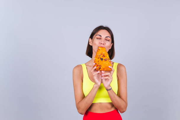 Sports woman stands on gray background, satisfied with the results of fitness training and diet, enjoying  piece of delicious pizza - Fotoğraf, Görsel