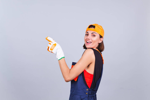 Malerin Arbeiterin in Overalls und Handschuhen auf grauem Hintergrund aufgeregt fröhliches Lächeln zeigen mit dem Finger nach links - Foto, Bild