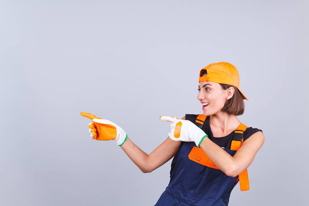 Painter worker woman in overalls and gloves on gray background excited cheerful smile point left with finger - Photo, Image