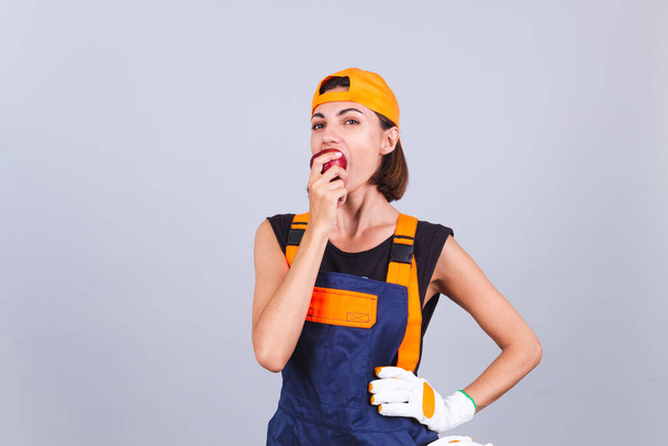 Pintor mujer trabajadora en overoles y guantes sobre fondo gris feliz disfrutando de manzana en el descanso - Foto, Imagen