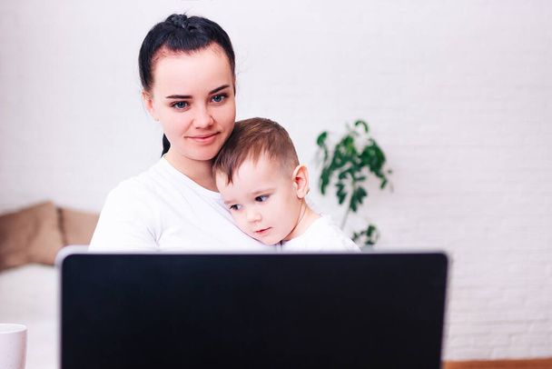 μια νεαρή μαυρομάλλα μητέρα με ένα μικρό παιδί εργάζεται στο σπίτι σε ένα φωτεινό δωμάτιο με ένα φορητό υπολογιστή - Φωτογραφία, εικόνα