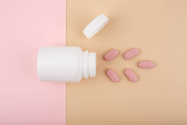 White opened medication bottle with pink spilled pills against pastel pink and beige background - Fotó, kép