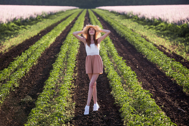 Young woman in white T-shirt, powdery shorts runs along green plowed field. Long hair develops at sunset, the girl has straw hat. Promote agricultural perspective, the texture of black soil ground - Φωτογραφία, εικόνα