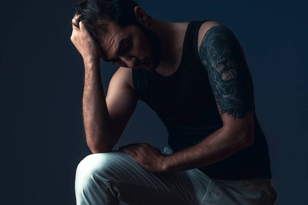 Man in studio on dark background. Guy with tattoo sits holding head. Concept of problem, frustration, concern - Foto, imagen