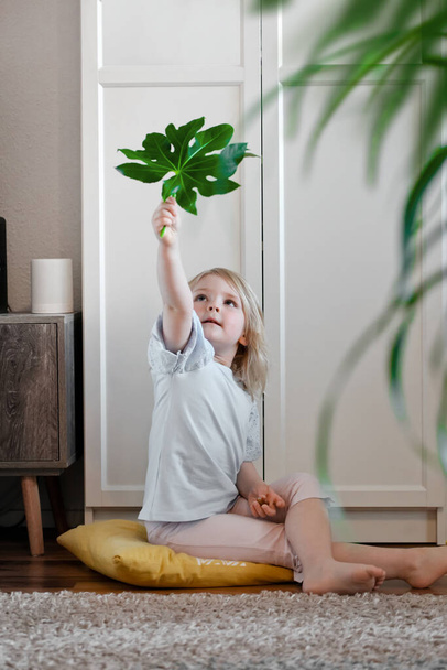 Маленька дівчинка сидить на підлозі у вітальні і грає з екзотичним листям рослин
 - Фото, зображення
