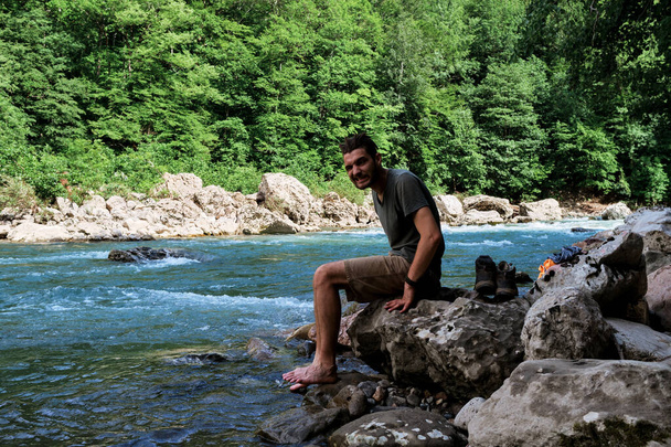 Ο άνθρωπος είναι τουρίστας. Νεαρός όμορφος Καυκάσιος Ταξιδιώτης με κοτσίδες και γένια πλένει τα πόδια του στο παγωμένο ποτάμι του βουνού και κάθεται πάνω στο βράχο. Βρέχει πόδια στο παγωμένο ποτάμι. - Φωτογραφία, εικόνα