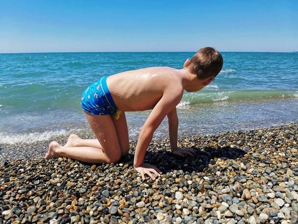 ένα αγόρι με μαγιό κάθεται στην ακτή της Μαύρης Θάλασσας μετά το κολύμπι - Φωτογραφία, εικόνα