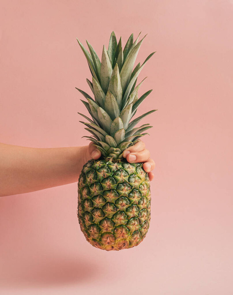 Creative exotic fruit idea with a woman's hand holding fresh organic pineapple. Minimalistic summer vibes concept, pastel pink background. Healthy food vertical arrangement. - Фото, изображение