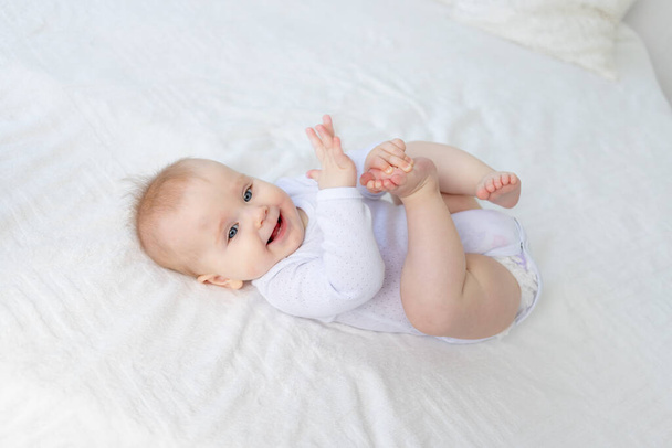 bébé fille souriante de six mois joue avec ses jambes couchées sur le dos sur un lit en coton blanc dans la chambre de la maison et rit. - Photo, image