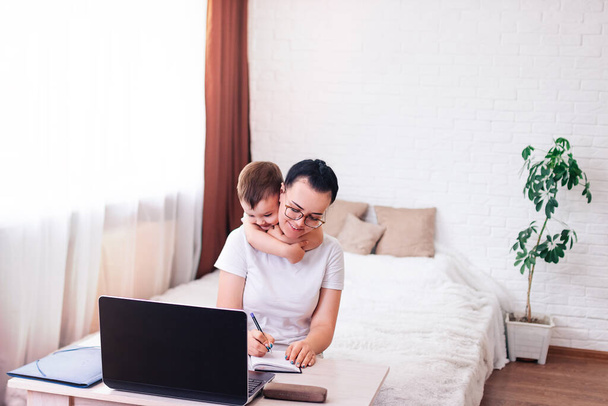 uma jovem mãe de cabelos pretos com uma criança pequena trabalha em casa em um quarto brilhante com um laptop - Foto, Imagem