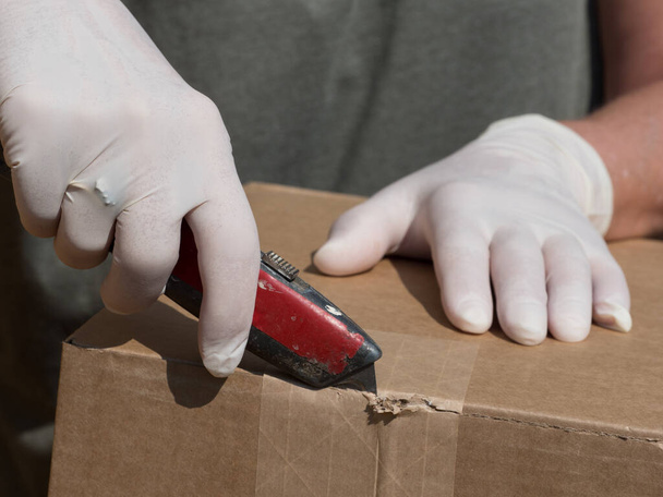 Une main dans un gant médical en latex blanc utilise un couteau artisanal pour couper le ruban adhésif.Une autre main gantée tient l'emballage comme mesure de protection contre le coronavirus. - Photo, image